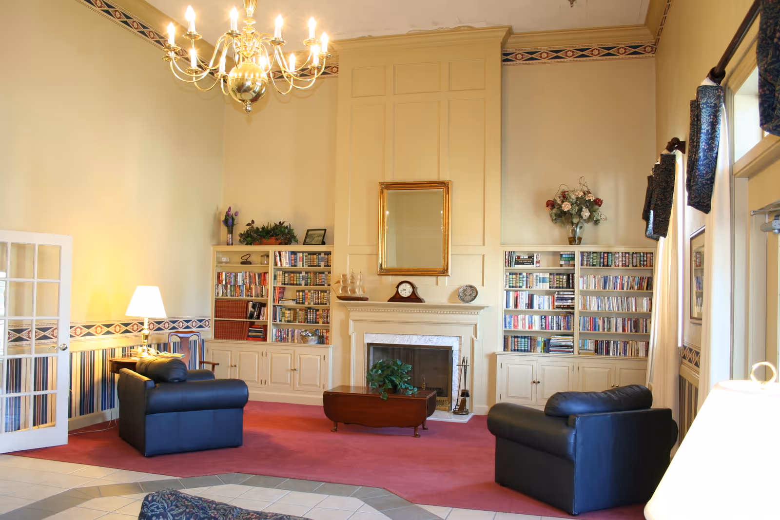 A cozy living room area with two black leather armchairs facing a fireplace. The fireplace is flanked by built-in bookshelves filled with books and decorative items. Above the fireplace is a large mirror with a gold frame. The room has high ceilings with a chandelier, patterned wallpaper border, and a red carpet. There are windows with dark curtains on the right side and a glass-paneled door on the left.