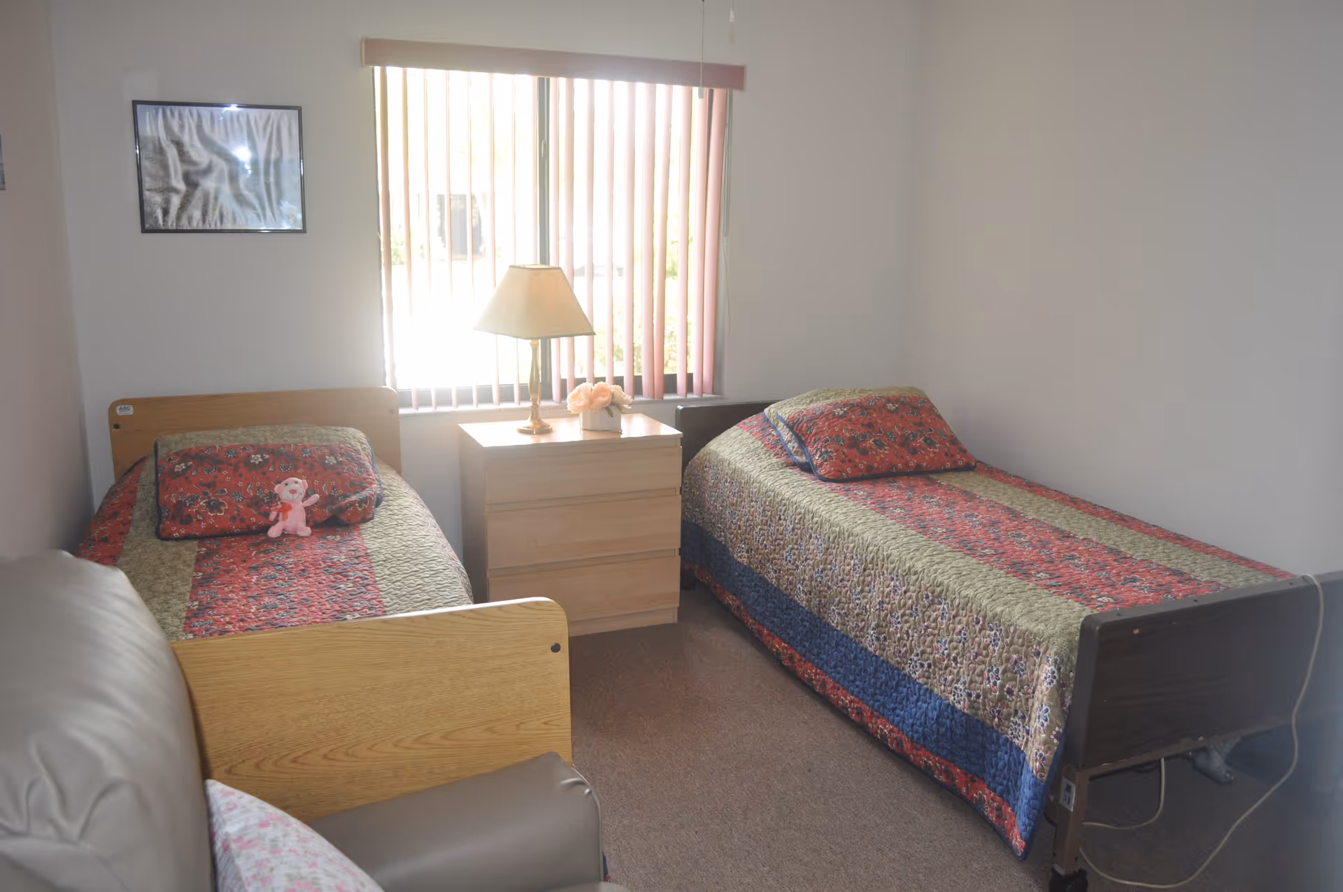 A small bedroom with two single beds, each covered with a patterned quilt in red, green, and blue tones. A nightstand with a lamp and a small flower arrangement is positioned between the beds. A window with vertical blinds is behind the nightstand, and a framed picture hangs on the wall above one bed. A beige armchair is partially visible in the foreground.