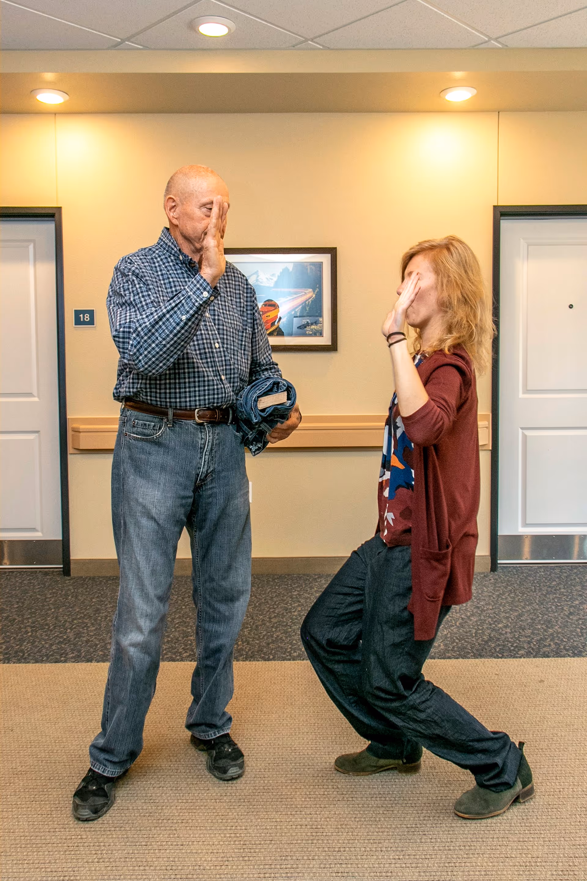 An elderly man and a woman standing in a hallway of a senior living facility, giving each other a high five. The man is holding a folded denim jacket and both are smiling. The hallway has beige walls, two white doors, a framed picture, and a room number 18 sign.