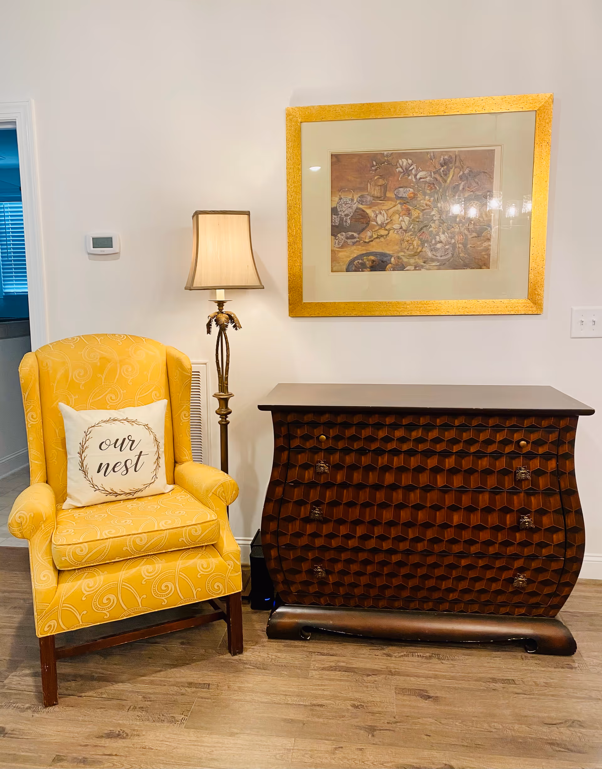 A cozy corner featuring a yellow upholstered armchair with a white pillow that reads 'our nest', a tall floor lamp with a beige shade, a dark wooden chest of drawers with a geometric pattern, and a framed artwork with a gold frame hanging on a white wall.