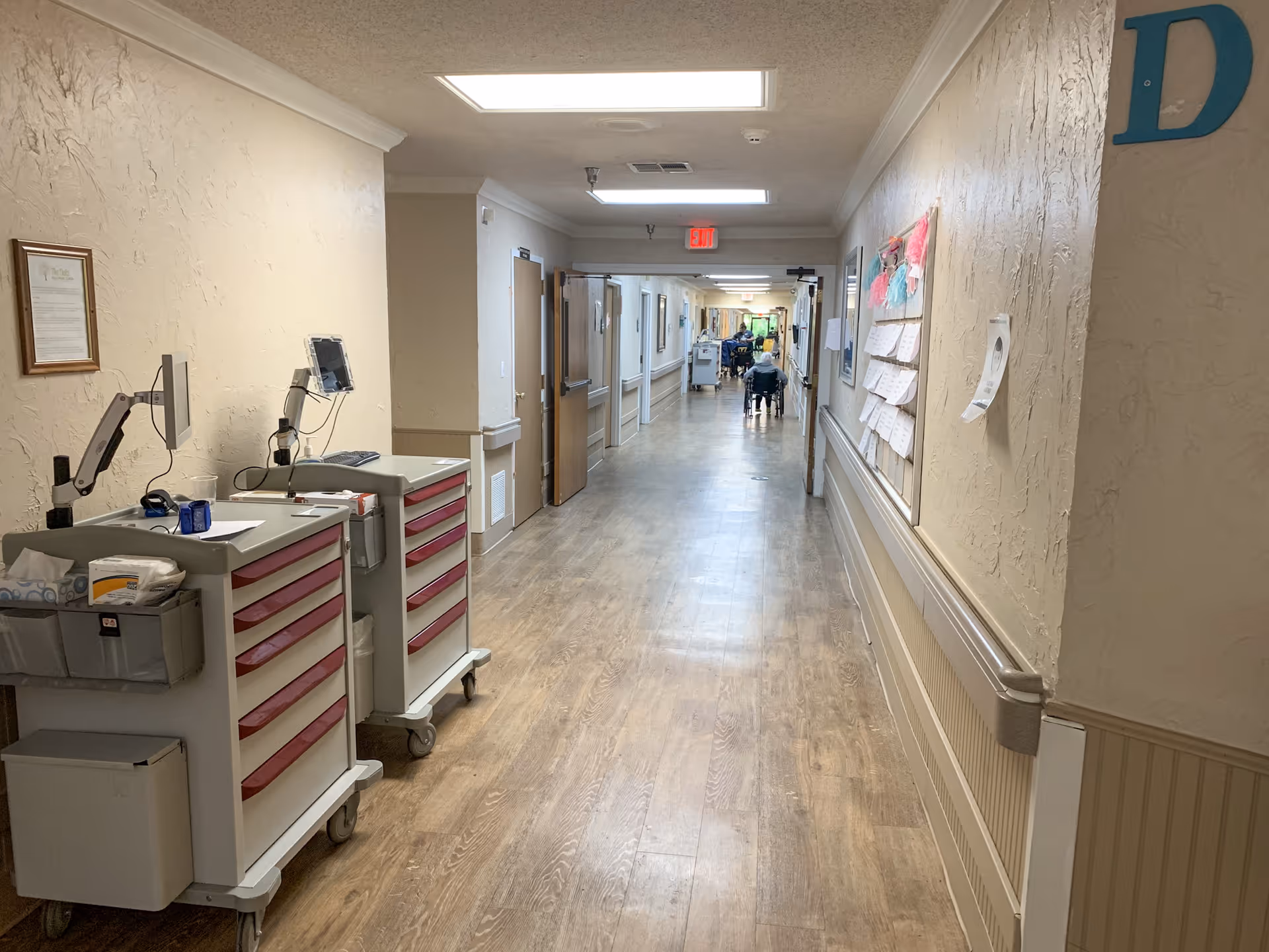 A long hallway in a healthcare facility with wooden flooring and beige textured walls. On the left side, there are two medical carts with monitors and supplies. The hallway has handrails on both sides and a bulletin board with papers pinned on the right wall. At the far end, a person in a wheelchair is visible moving down the corridor. The ceiling has fluorescent lighting and an exit sign is visible above a door.
