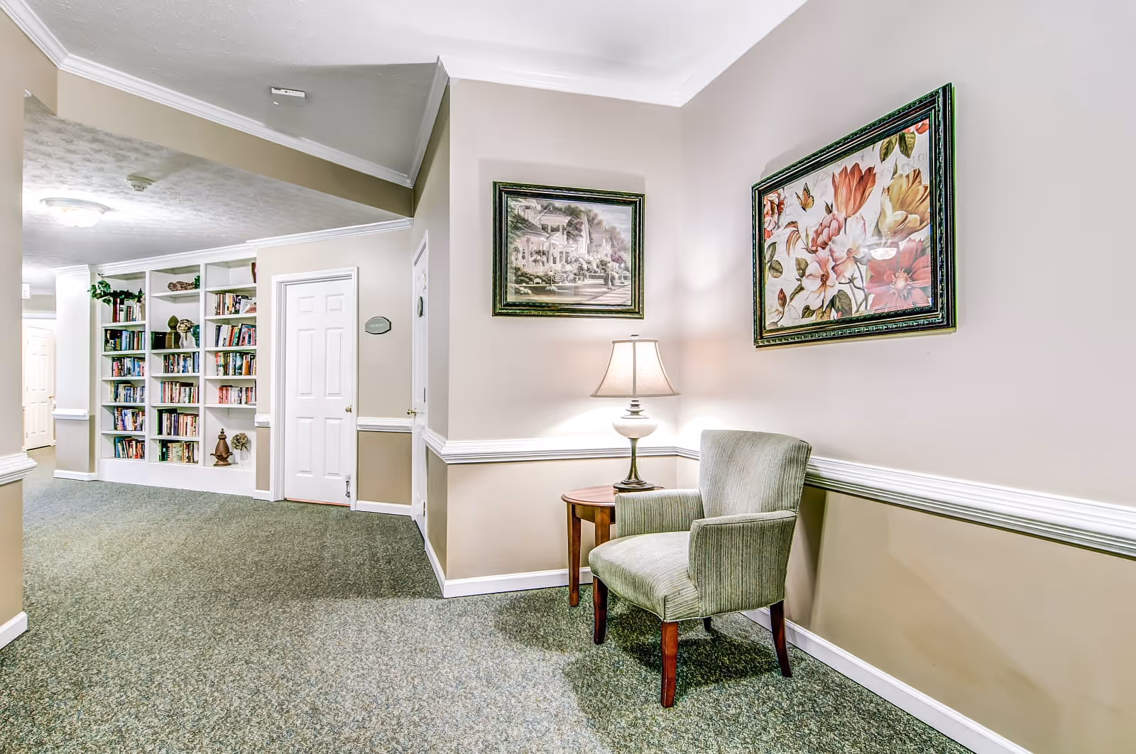 A hallway corner in a senior living facility with beige walls and green carpet. There is a green upholstered armchair next to a small wooden side table with a lamp. Two framed paintings hang on the walls above the chair and table. In the background, there is a built-in white bookshelf filled with books and decorative items, and two closed white doors.