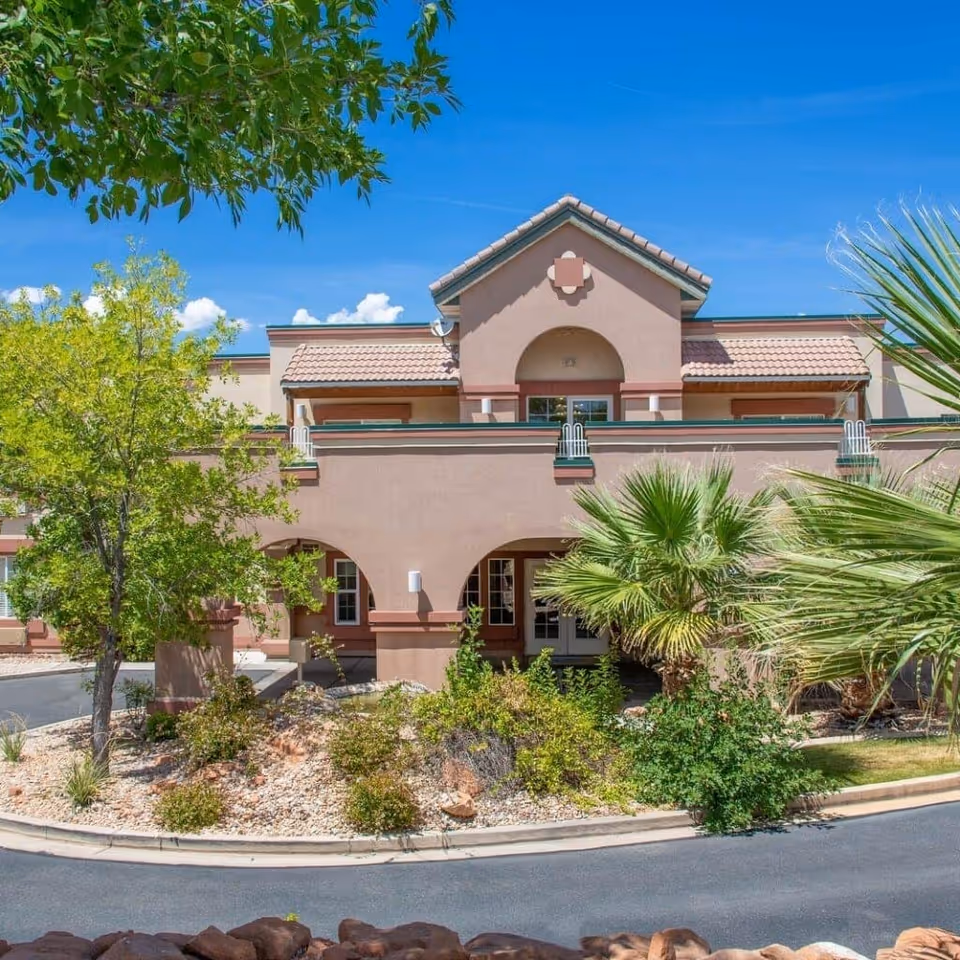 Front exterior of a two-story stucco senior living building with arched entryways, palm trees, and landscaped rock beds under a blue sky.