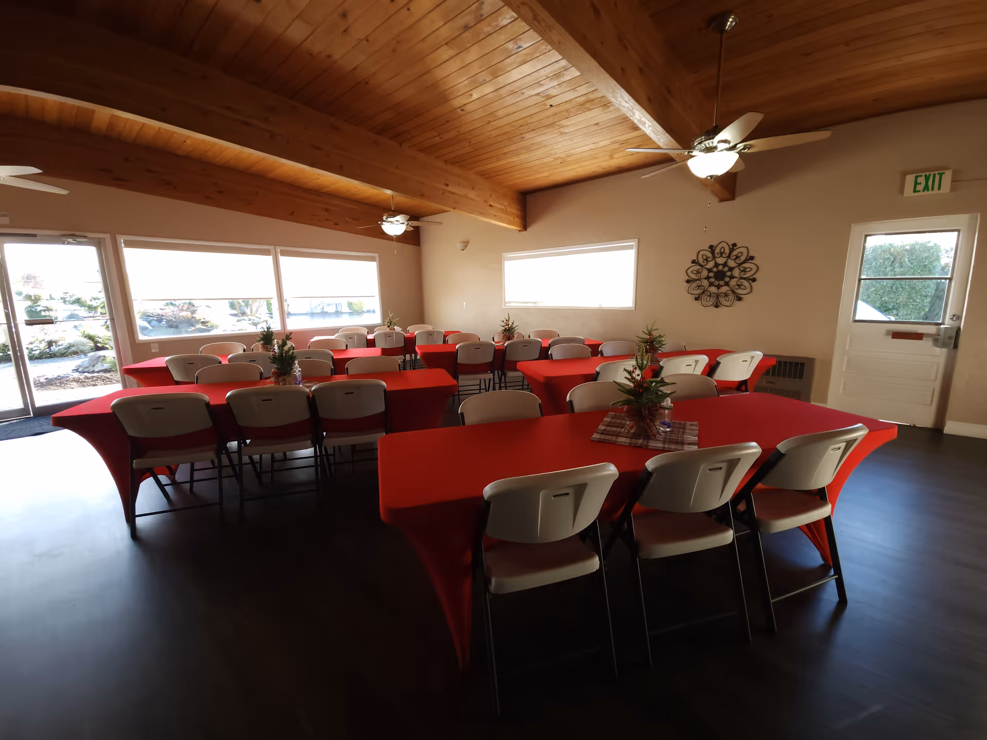 Interior of a community dining room with several tables covered in red tablecloths and white chairs arranged around them. The room has a wooden ceiling with exposed beams and ceiling fans with lights. There are large windows letting in natural light, a white door with an exit sign above it, and small decorative plants on the tables.