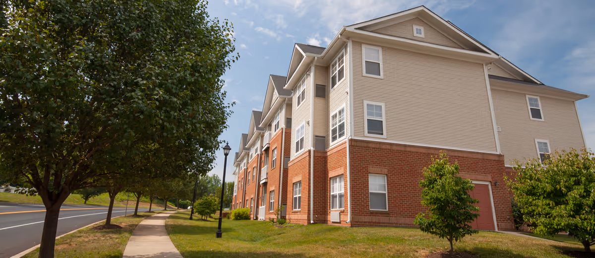 Exterior view of a multi-story senior living facility building with beige siding and red brick lower walls, surrounded by green grass, small trees, and a sidewalk along a street under a partly cloudy sky.