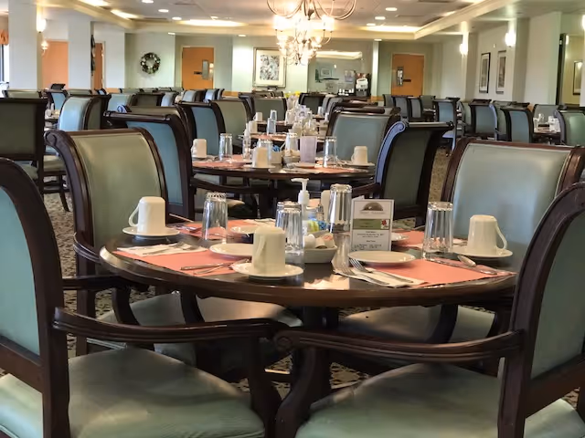 Dining room with multiple round tables set for meals and green upholstered chairs.