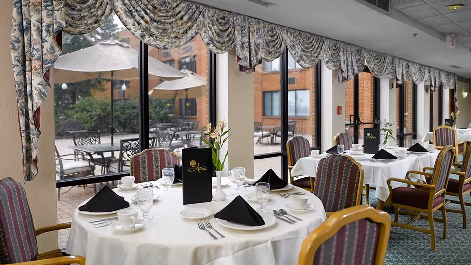 Dining room with round and rectangular tables covered with white tablecloths, set with black napkins, glassware, and white dishes. The room has large windows with floral valances, offering a view of an outdoor patio with tables and umbrellas. Chairs have wooden arms and striped upholstery.