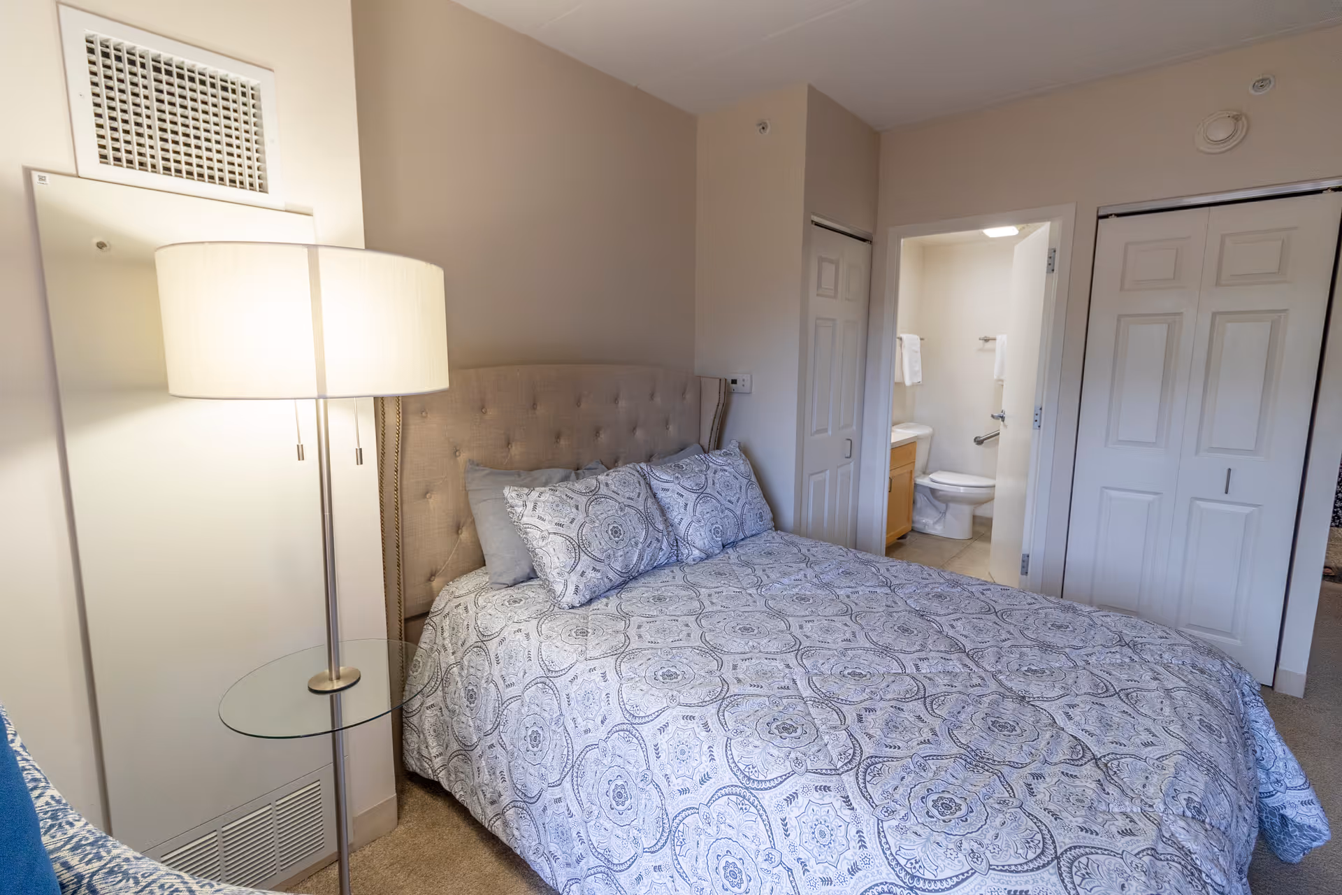 A bedroom with a bed covered in a patterned blue and white comforter and matching pillows. To the left of the bed is a floor lamp with a glass side table. In the background, there is a doorway leading to a bathroom with a toilet and a vanity with a sink. There are also two closed white closet doors on the right side of the image.