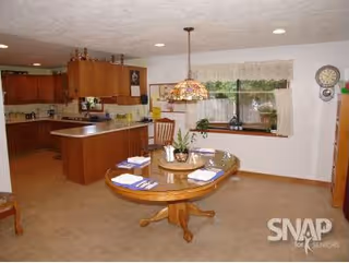 A spacious kitchen and dining area with wooden cabinets and a central island in the kitchen. In the foreground, there is a round wooden dining table set with placemats, napkins, and a centerpiece. A decorative hanging lamp is above the table. A large window with curtains lets in natural light, and a wall clock is visible on the right side.