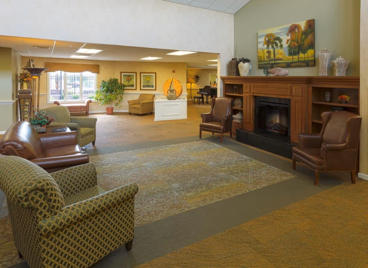 A spacious senior living facility common area with comfortable seating including patterned armchairs and leather chairs arranged around a wooden fireplace. The room features a large decorative rug, a painting of trees above the fireplace, and various decorative vases. In the background, there is a window with a bench, potted plants, framed artwork on the walls, and a piano in an adjoining room.