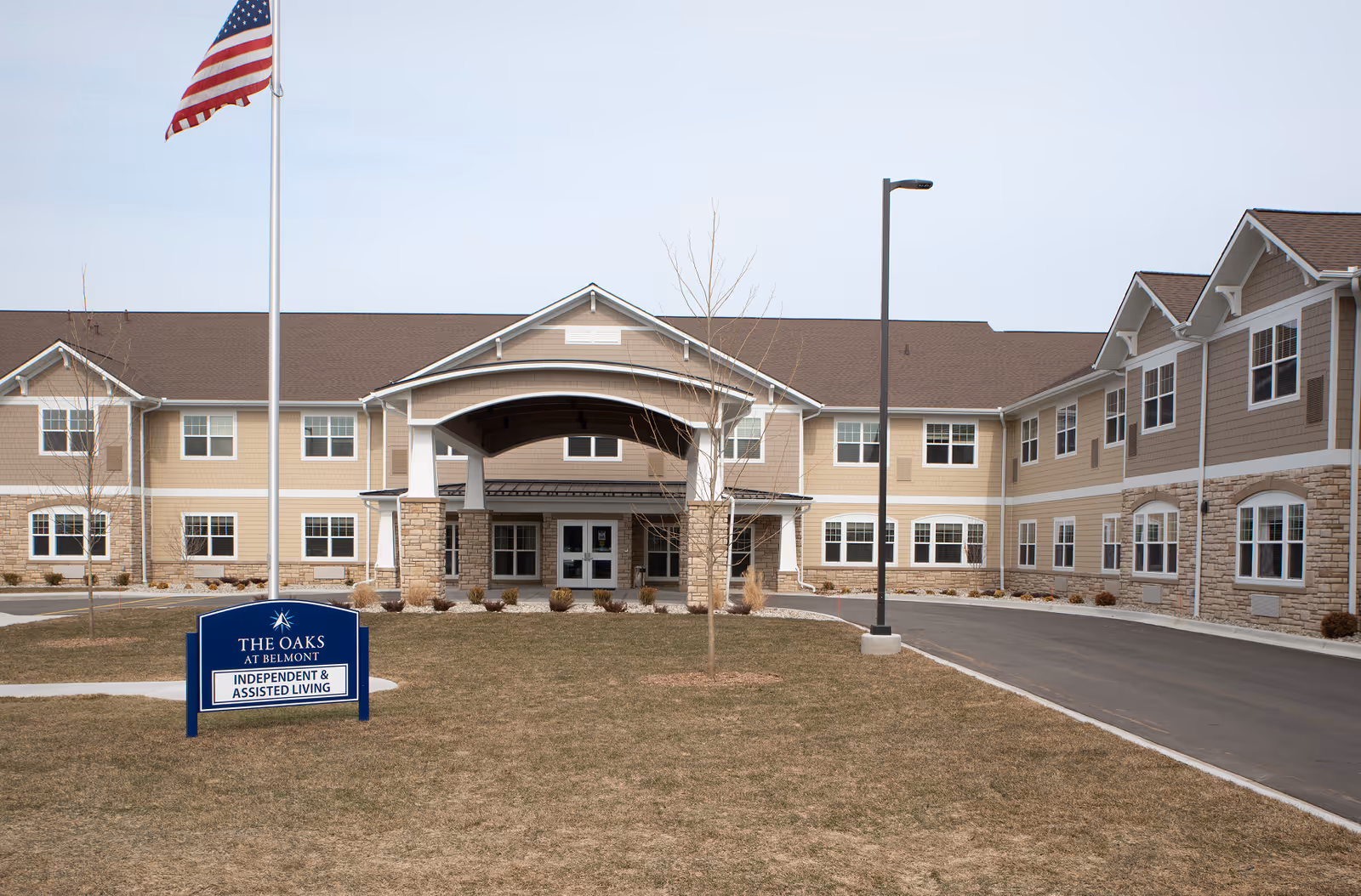 Exterior view of The Oaks at Belmont senior living facility showing a two-story building with beige siding and stone accents. There is a covered entrance with pillars, an American flag on a flagpole, a street lamp, and a sign in front that reads 'The Oaks at Belmont Independent & Assisted Living'. The sky is clear and the grass is brown.