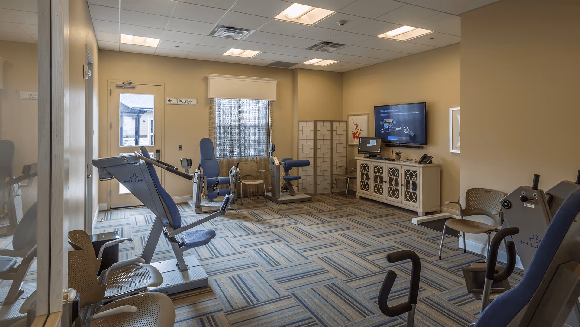 A well-lit exercise room with various fitness equipment including weight machines and chairs. The room has patterned carpet flooring, beige walls, a window with a curtain, a door, and a TV mounted on the wall above a white cabinet. There are also some framed pictures and a folding screen in the corner.