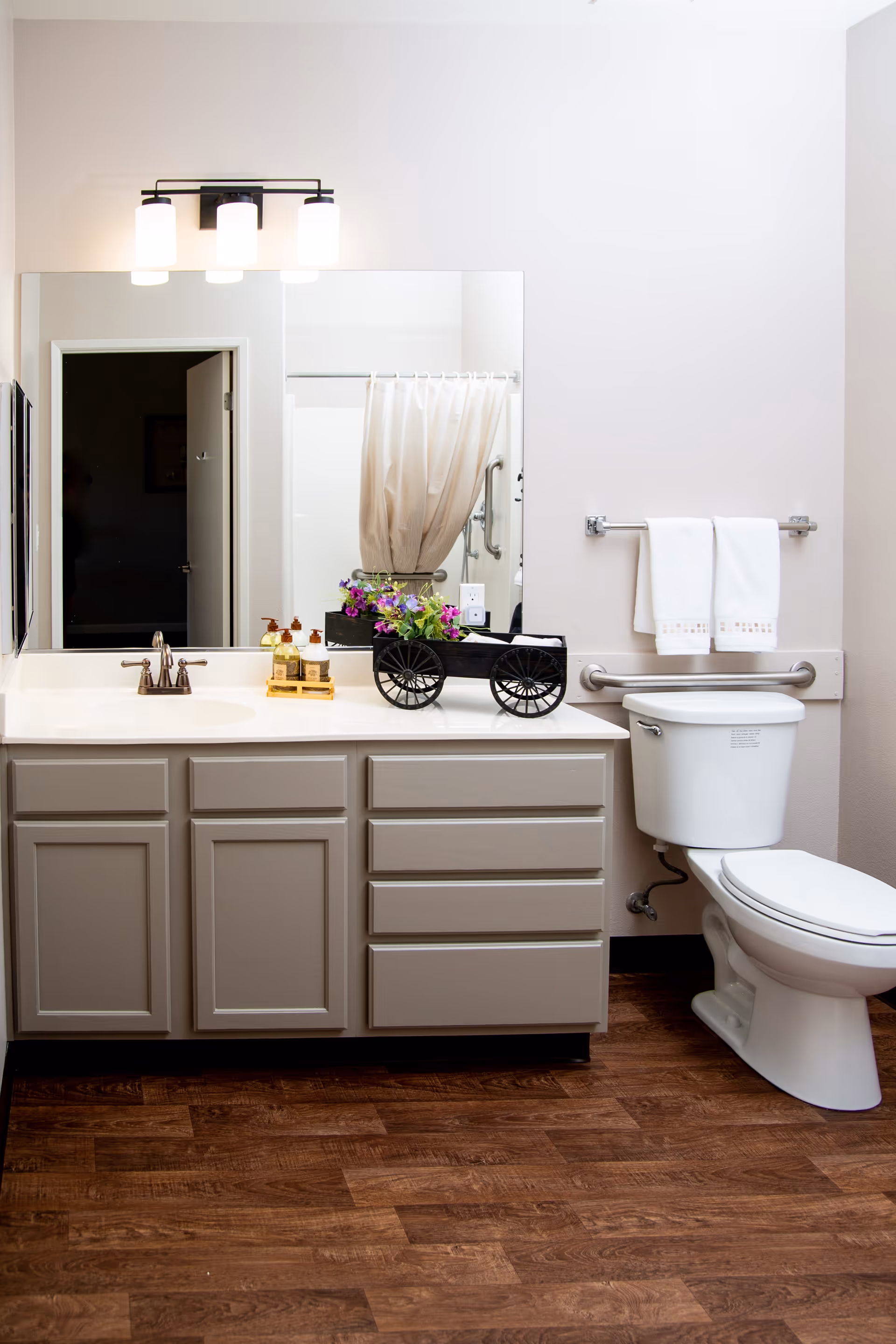A clean and well-lit bathroom featuring a white toilet with grab bars, a vanity with a sink, a large mirror above the sink, and a small decorative black cart with purple flowers on the countertop. Two white towels hang on a towel rack above the toilet. The floor has a wood-like finish.