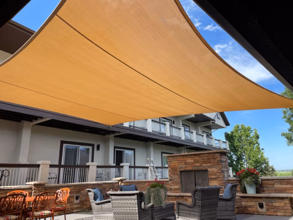 Outdoor patio seating area with wicker chairs, a stone fireplace, dining chairs, and a large tan sun sail in front of a multi-story building.