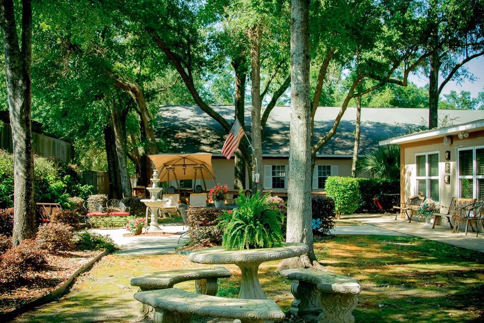 Outdoor garden area at Grandview Retirement Center featuring a stone table with curved benches, surrounded by trees and shrubs. In the background, there is a patio with chairs, a fountain, and an American flag hanging from a tree. The area is shaded and appears peaceful and well-maintained.