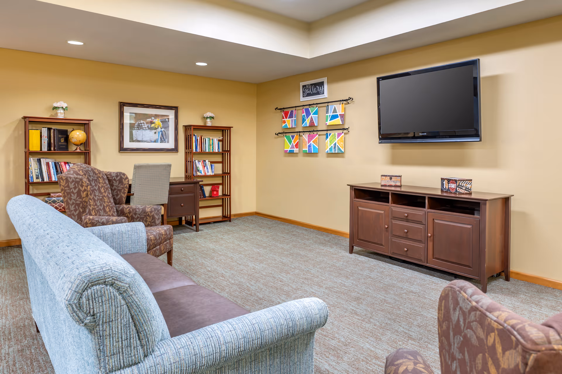 A cozy living room area with a light blue sofa, two patterned armchairs, a wooden TV stand with a flat-screen TV mounted on the wall above it, two wooden bookshelves filled with books and decorative items, a small desk with a chair, and colorful abstract artwork on the wall labeled 'Art Gallery'.