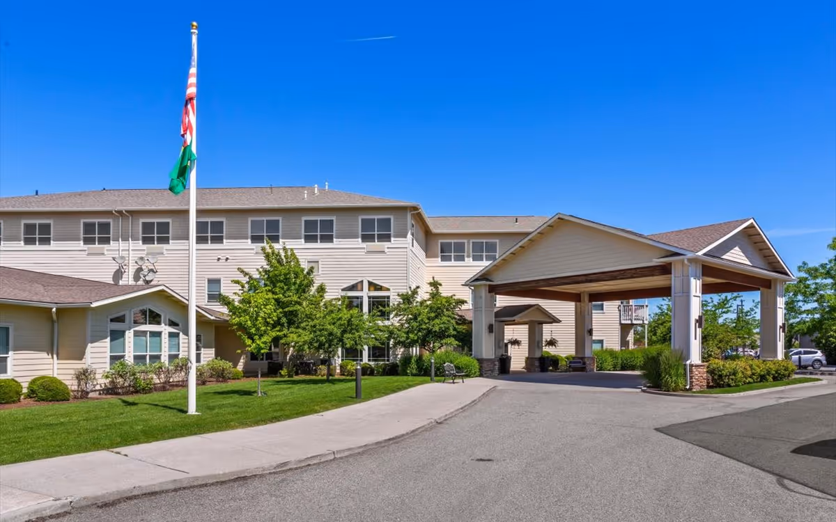 Exterior view of Vineyard Park of South Hill, showing a large beige building with multiple windows, a covered entrance with pillars, a flagpole with American and green flags, and landscaped greenery under a clear blue sky.