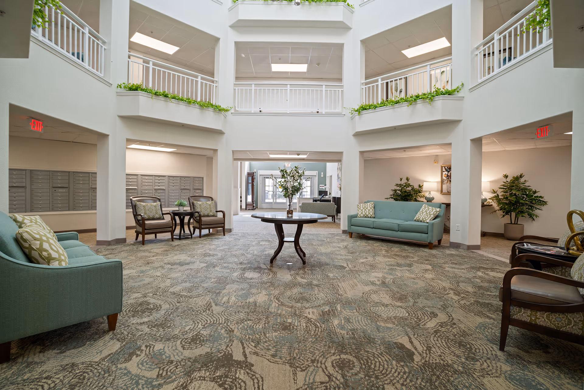 Spacious and well-lit interior common area with high ceilings and a second-floor balcony. The room features two teal sofas with patterned pillows, several armchairs, a round wooden table with a floral arrangement in the center, and potted plants. Mailboxes are visible on the left wall, and exit signs are illuminated above doorways on both sides.