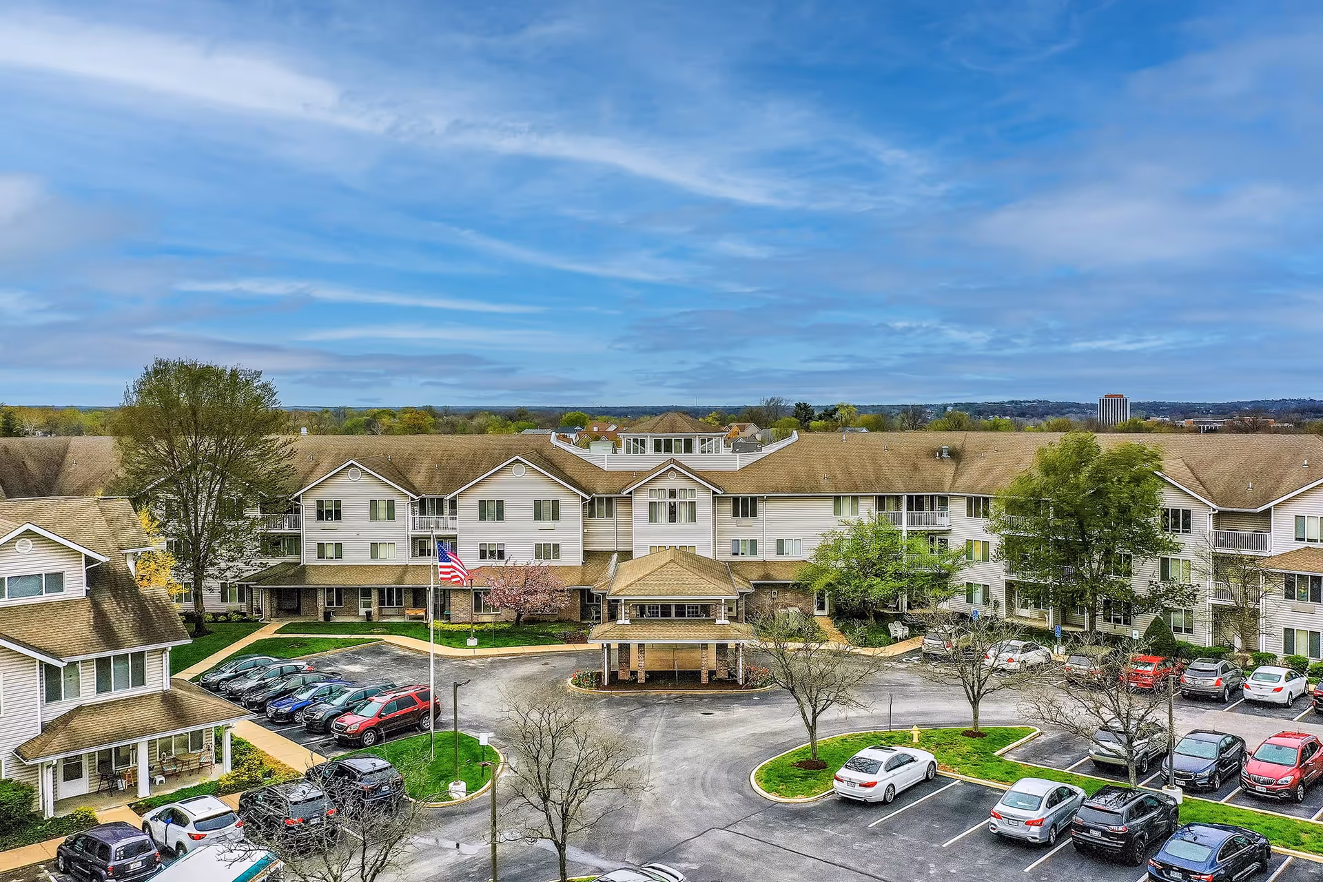 Front exterior of a three-story senior living building with a covered entrance, circular driveway, landscaped grounds, and parked cars under a blue sky.