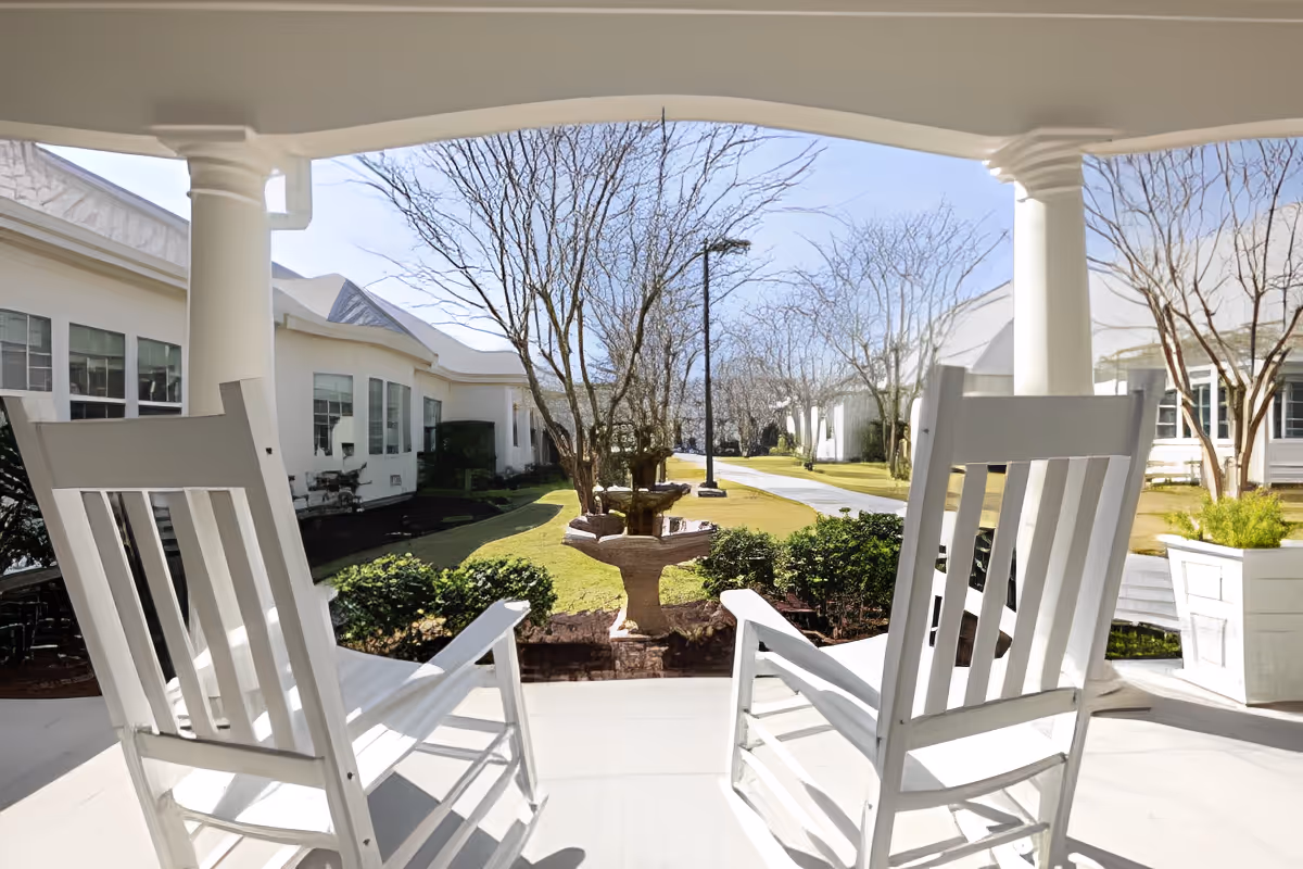 View from a covered porch with two white wooden rocking chairs facing a landscaped garden area with a birdbath, trees without leaves, and a paved walkway leading between white buildings under a clear blue sky.