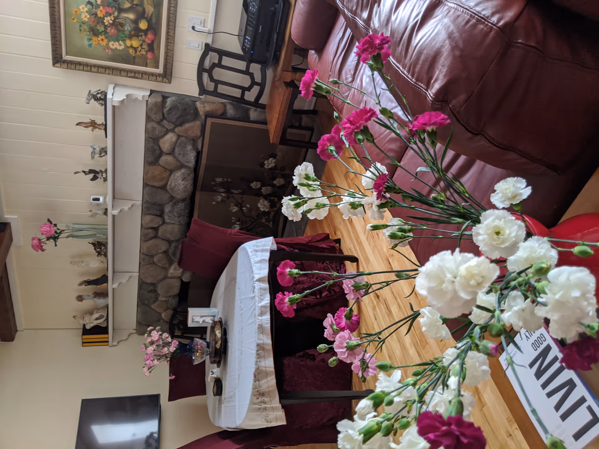A cozy living room with a stone fireplace, a round table covered with a white tablecloth and surrounded by chairs with red covers. In the foreground, there is a vase with white and pink flowers on a wooden surface. The room has wooden floors, a painting on the wall above the fireplace, and a TV mounted on the wall.