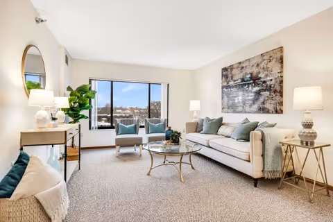Bright living room with a sofa, two armchairs by a large window, a round glass coffee table, and decorative lamps and artwork.