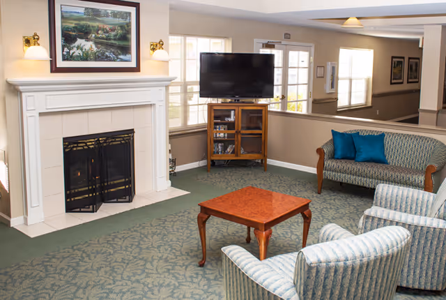 A cozy living room area with a fireplace on the left, a wooden coffee table in the center, two striped armchairs in the foreground, and a patterned sofa with blue cushions against the wall. A flat-screen TV is placed on a wooden stand near windows that let in natural light.