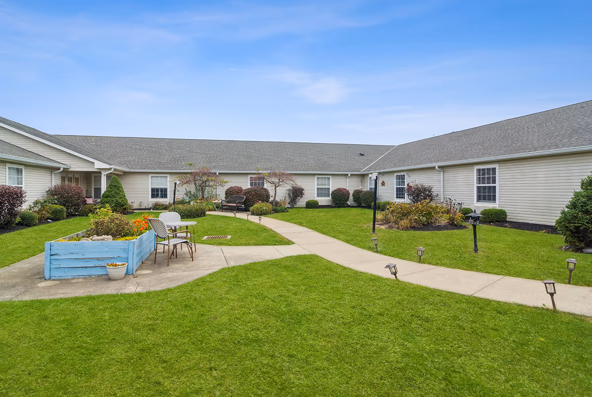 A well-maintained grassy courtyard with paved walkways, a raised planter and chairs surrounded by single-story ranch-style buildings under a blue sky.