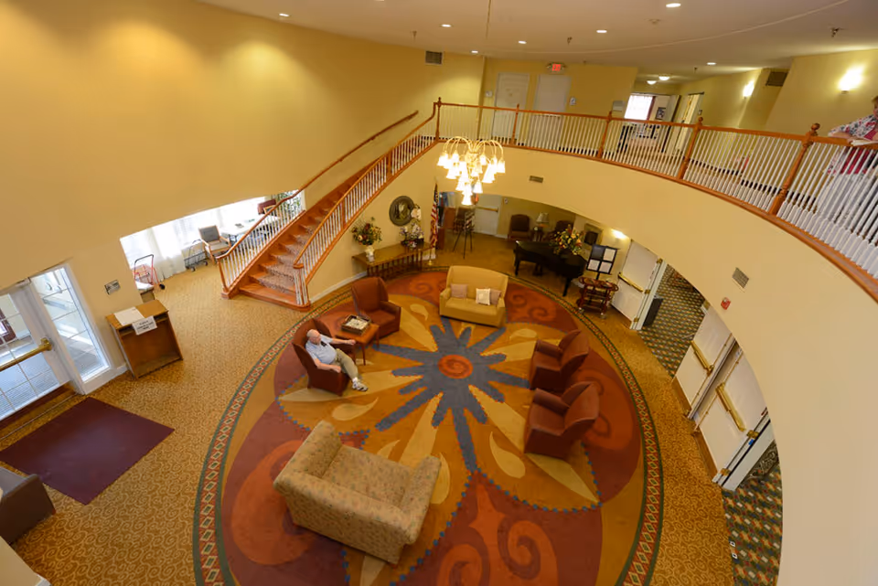 A spacious senior living facility common area with a large circular patterned carpet in the center, surrounded by various chairs and sofas. A man is seated in one of the chairs. There is a staircase with wooden railings leading to an upper level balcony overlooking the area. The room is well-lit with ceiling lights and a chandelier hanging above the seating area. The walls are painted a light yellow, and there are doors and windows visible around the room.
