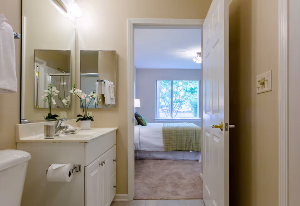 View from a bathroom looking into a bedroom. The bathroom has a white vanity with a sink, a mirror, a towel rack with a white towel, and a toilet with a toilet paper holder. The bedroom has a bed with white bedding and a green quilt, a window showing trees outside, and a lamp on a bedside table.