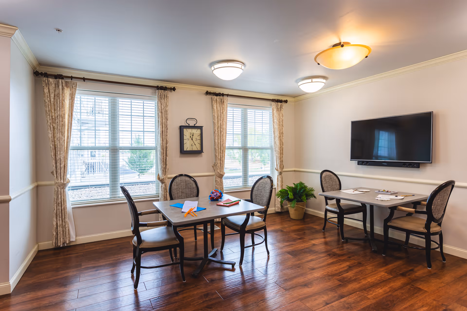 A well-lit room with two tables and six chairs arranged around them. The room has large windows with beige curtains, a wall clock between the windows, a flat-screen TV mounted on the wall, and a potted plant in the corner. The floor is wooden, and the ceiling has three light fixtures.