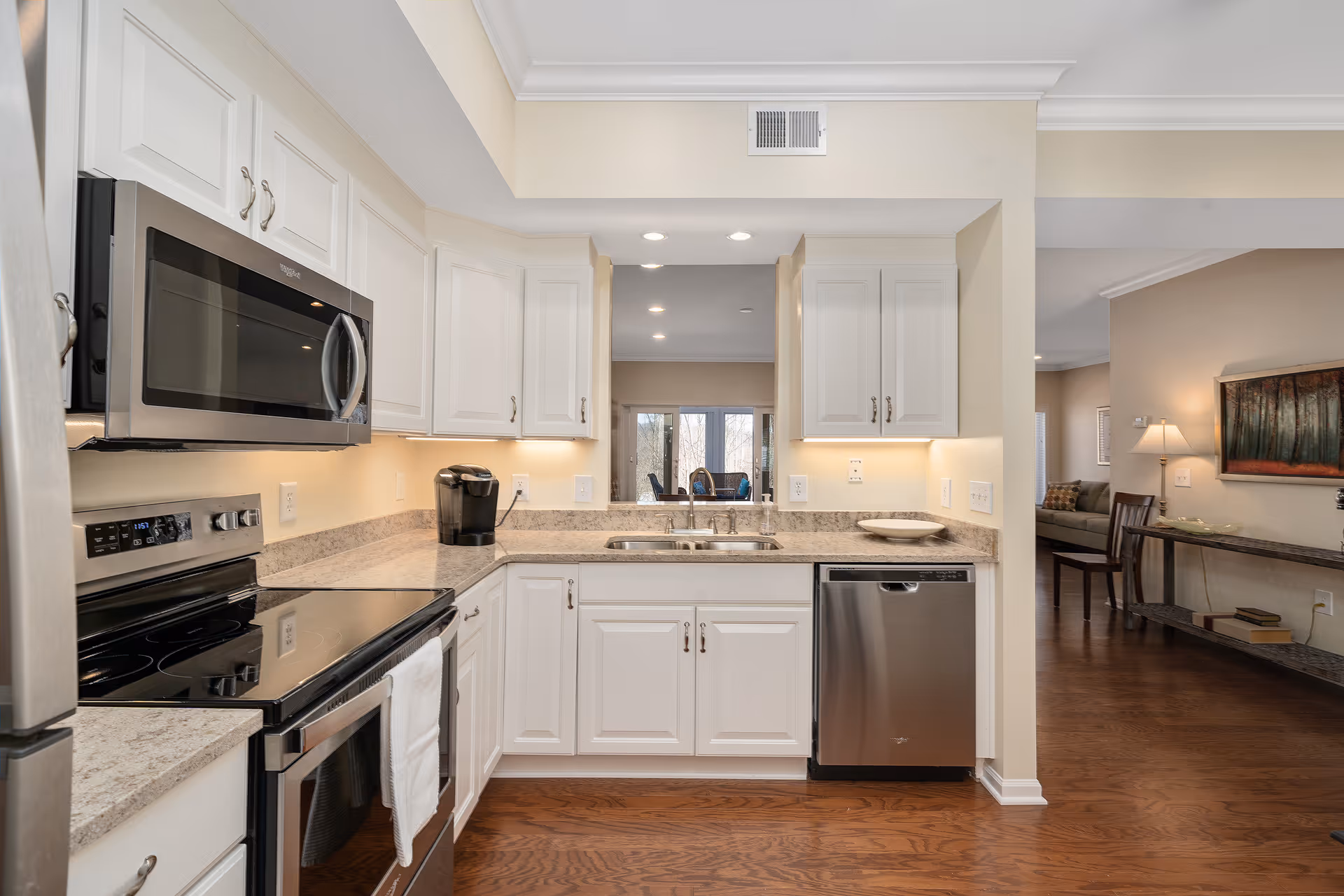 Modern kitchen with white cabinets, granite countertops, stainless steel appliances including a microwave, stove, and dishwasher. The kitchen has a double sink under a pass-through window opening to a living area with wooden floors and neutral-colored walls.