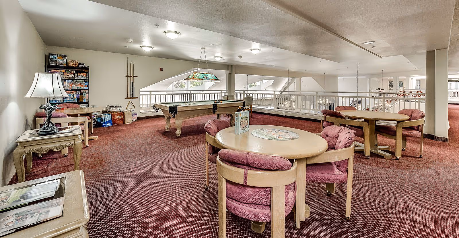 A spacious common area with red carpet flooring featuring two round wooden tables each surrounded by four cushioned chairs with pink upholstery. In the background, there is a pool table with a hanging light fixture above it. On the left side, there is a small table with a lamp and a shelving unit filled with board games and puzzles. The area is well-lit with ceiling lights and natural light coming through windows.