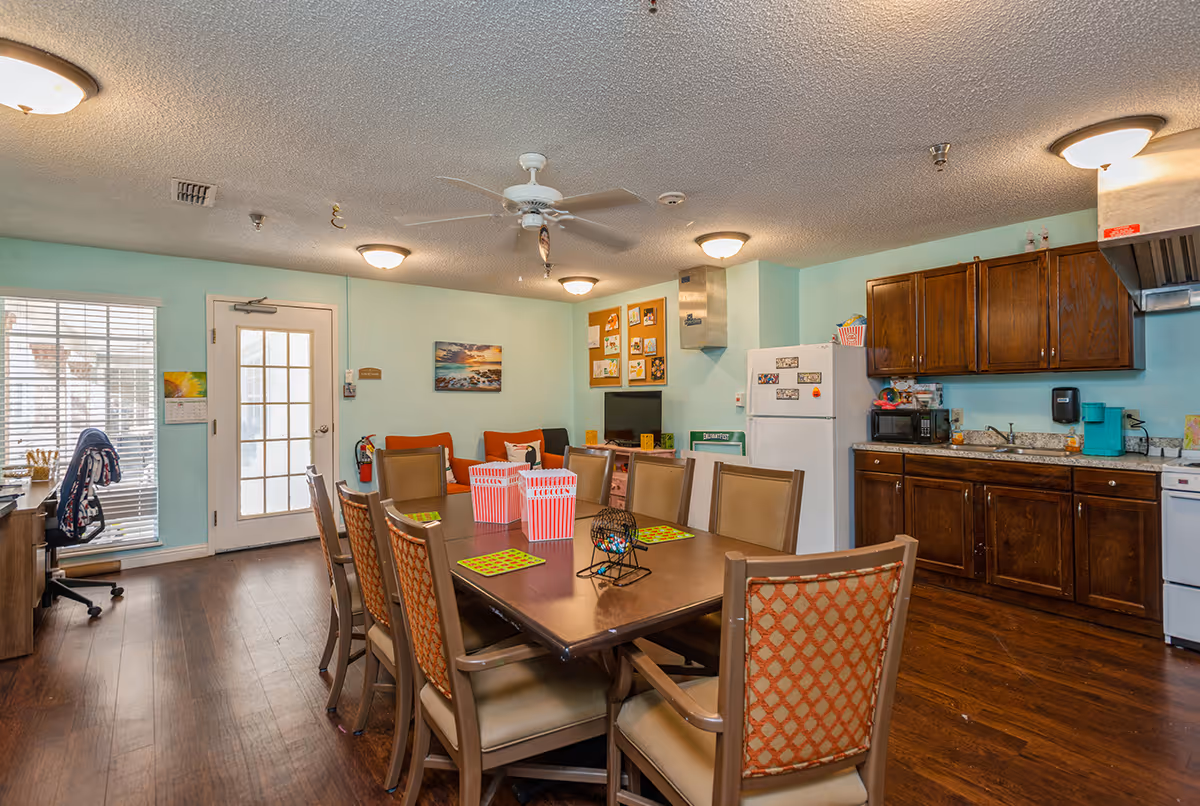 A communal room with a large wooden table surrounded by chairs, a kitchen area with wooden cabinets, a white refrigerator, microwave, and coffee maker. The room has light blue walls, a ceiling fan, and several ceiling lights. There is a door with glass panels and a window with blinds on the left side, and a small seating area with orange cushions and a TV in the background.