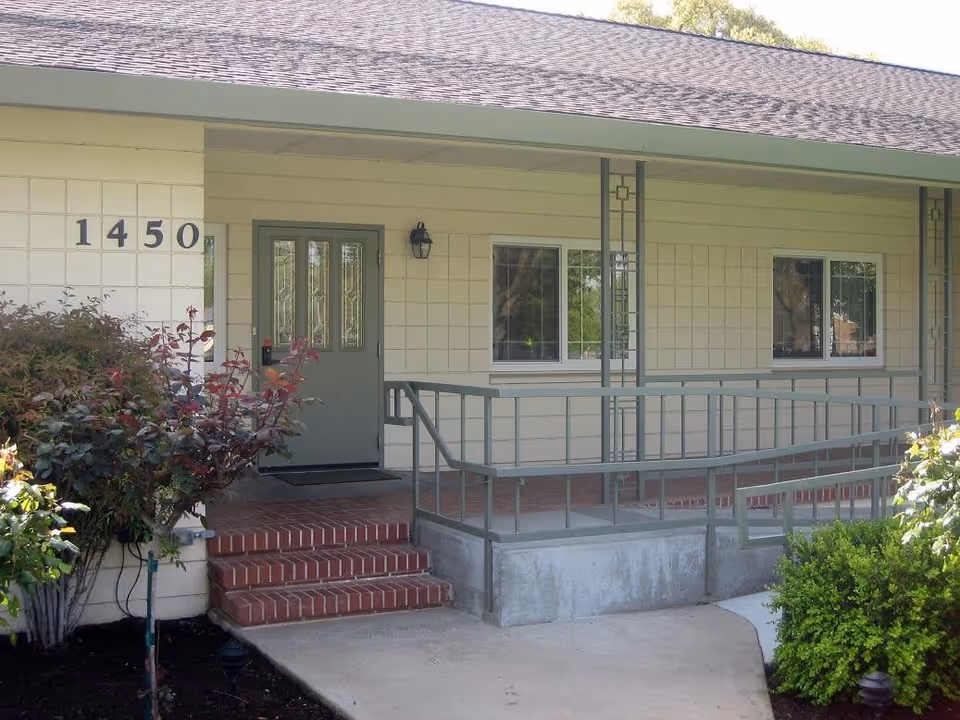 Front entrance of a single-story assisted living building with the number 1450, a ramp and brick steps leading to a glass-paneled door and windows.