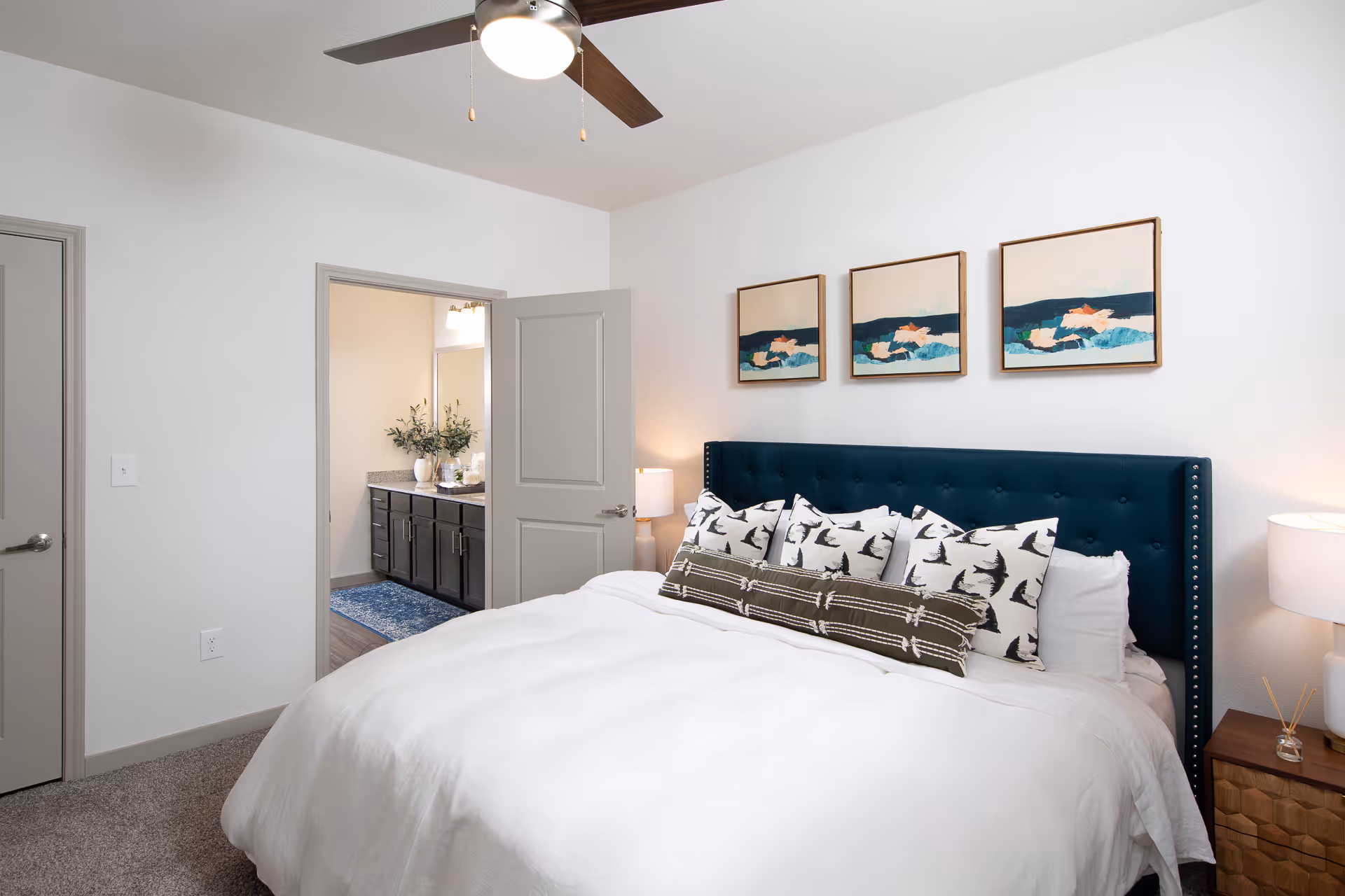 A bedroom with a large bed featuring a dark blue tufted headboard and multiple decorative pillows. Above the bed are three framed abstract paintings. To the right of the bed is a wooden nightstand with a white lamp and reed diffuser. The room has light-colored walls and carpeted flooring. An open door reveals a bathroom with a granite countertop, dark cabinets, a large mirror, and a blue rug.