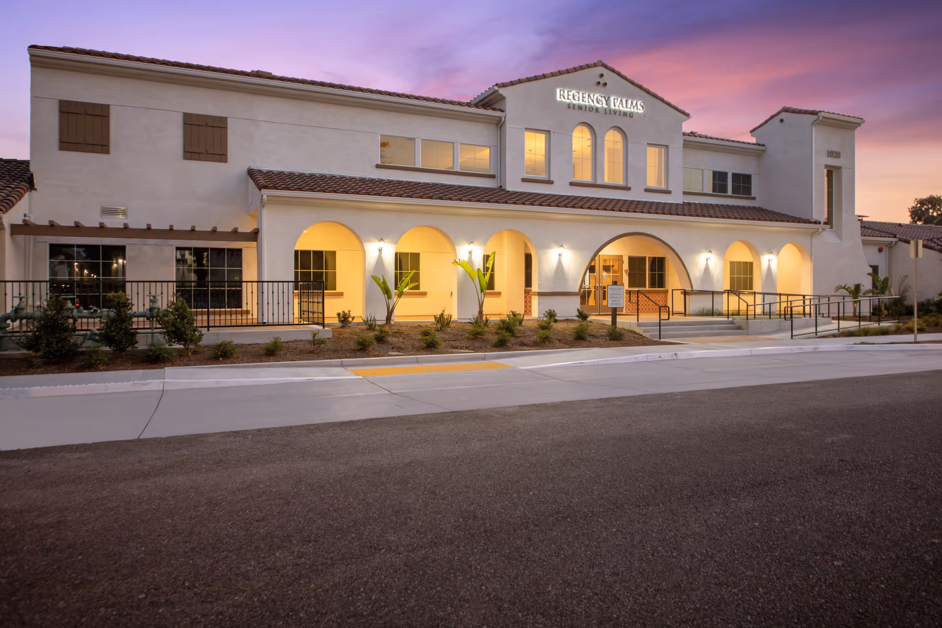 Exterior view of Regency Palms Senior Living facility at dusk, showing a two-story building with arched entryways, illuminated windows, and a landscaped front area with small plants and a paved driveway.