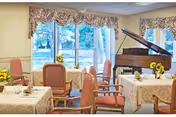 A dining room with tables covered in beige tablecloths, each set with napkins and small flower arrangements. There are several upholstered chairs around the tables. Large windows with floral valances let in natural light, and a grand piano is positioned near the windows.