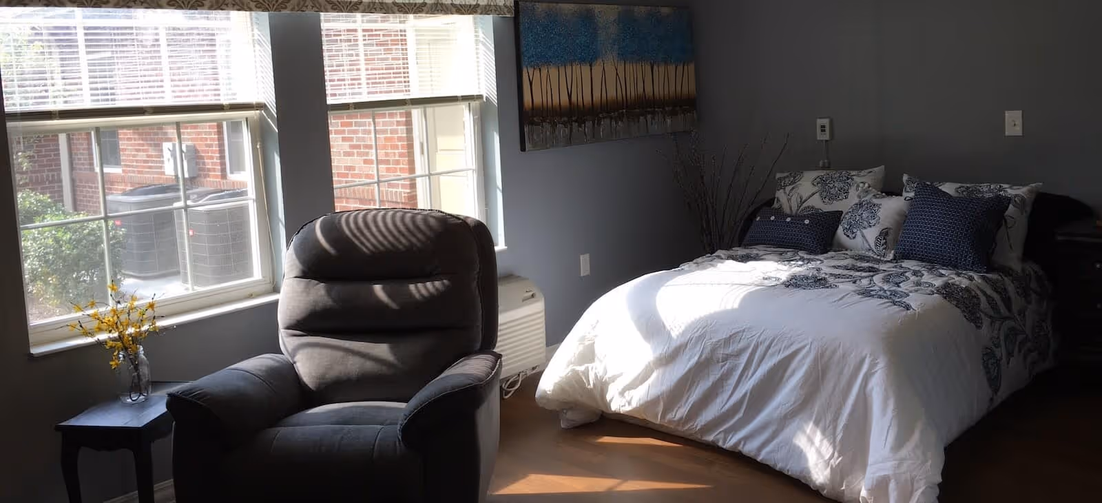 Sunlit bedroom with a bed, recliner chair, side table, and large windows.