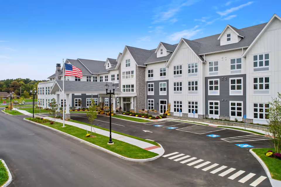 Exterior view of a large, modern senior living facility building with multiple windows and a covered entrance. An American flag is flying on a flagpole near the entrance. The parking lot in front has several handicap parking spaces and a pedestrian crosswalk. The sky is clear and blue.
