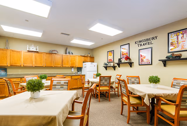 A dining area with multiple tables covered in beige tablecloths, each surrounded by wooden chairs with patterned cushions. The room has wooden cabinets along one wall, a white refrigerator, and a countertop. On the wall, there are framed travel posters and shelves with decorative items. The space is well-lit with ceiling lights and has a cozy, inviting atmosphere.