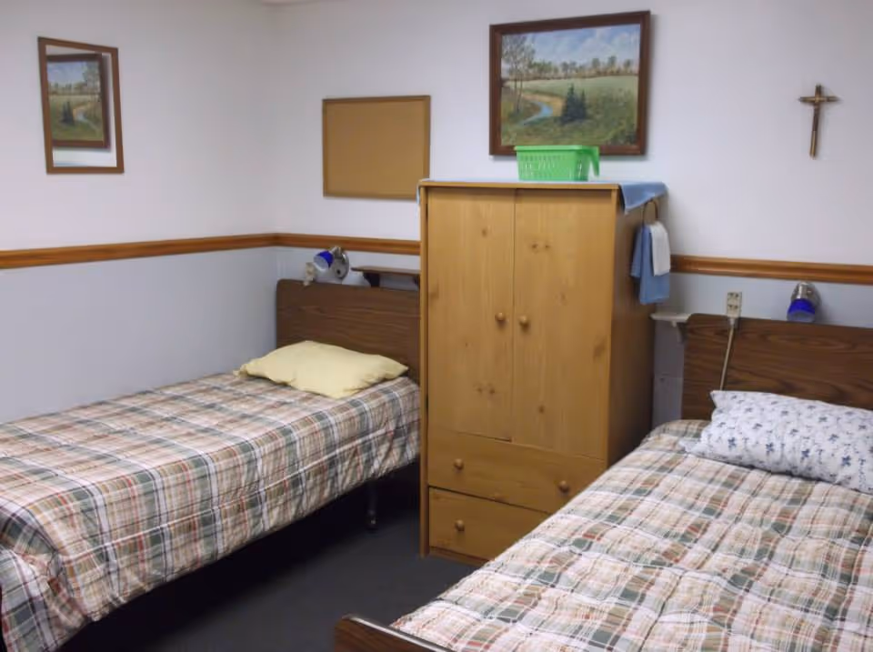A small bedroom with two single beds separated by a wooden wardrobe. Each bed has a plaid bedspread and a pillow. The walls are white with a wooden trim, and there are two framed landscape paintings and a crucifix hanging on the walls. Small reading lamps are attached to the headboards of each bed.