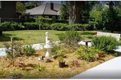A garden area with a small statue in the center surrounded by various plants and shrubs. There is a concrete pathway curving around the garden, and trees and a building are visible in the background.