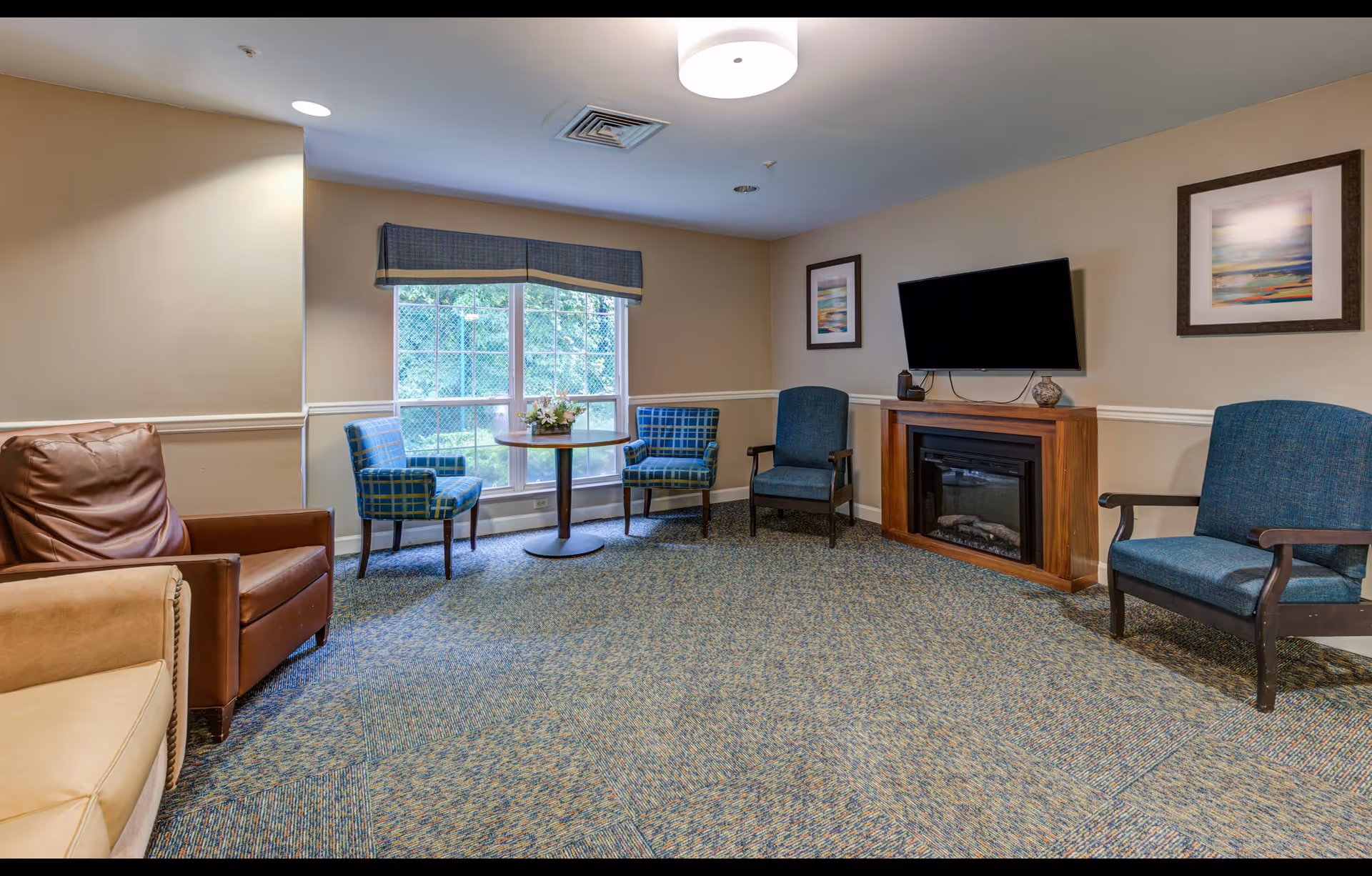 A cozy living room with a beige and blue color scheme featuring a large window with a blue valance, a round wooden table with a flower arrangement, four upholstered chairs in blue tones, a wall-mounted flat-screen TV above a wooden electric fireplace, and two framed landscape paintings on the wall.