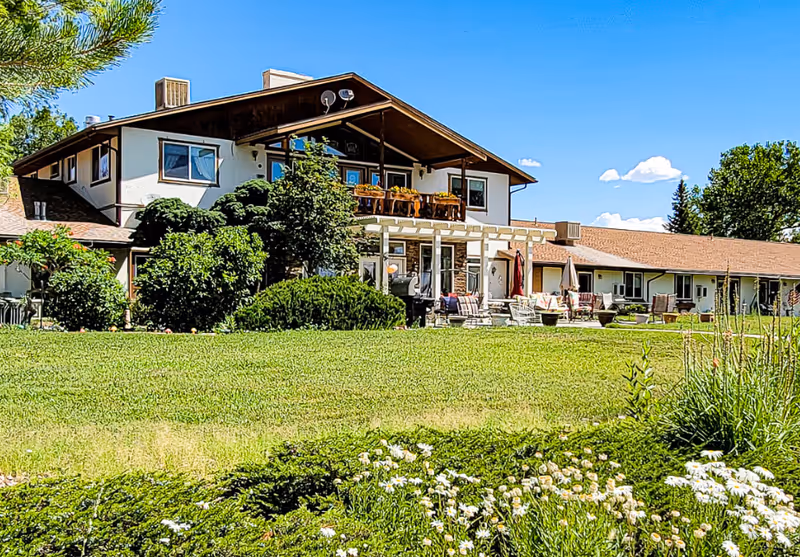 A large two-story building with a brown roof and white walls surrounded by green bushes and a well-maintained lawn. The building has a balcony with flower pots and a patio area with outdoor furniture. The sky is clear and blue with a few clouds.