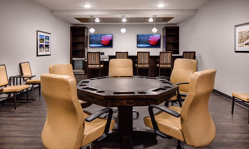 A well-lit room with a large octagonal table surrounded by six tan leather chairs with armrests. In the background, there are two computer monitors on a desk with four striped chairs. The room has wooden flooring, white walls, and framed pictures on the walls.