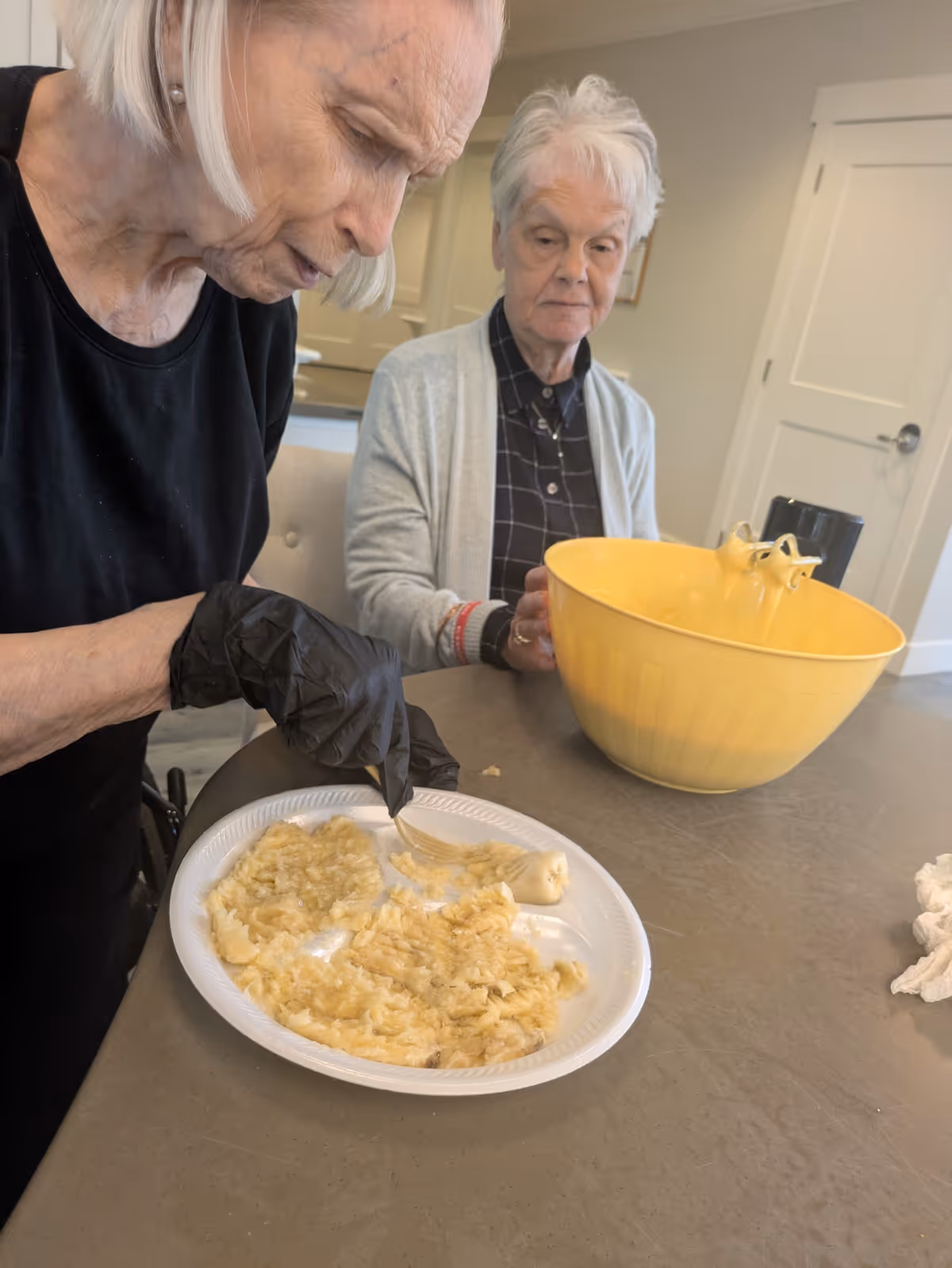 Two elderly women in a kitchen or dining area. One woman wearing a black shirt and black gloves is mashing bananas on a white disposable plate with a fork. The other woman, wearing a gray cardigan over a checkered shirt, is sitting and watching. A large yellow mixing bowl is on the table in front of them.