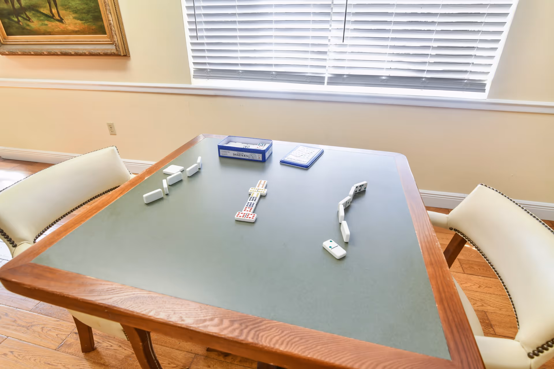 A wooden table with a green felt surface set up for a game of dominoes, with domino tiles arranged on the table and two cream-colored chairs with nailhead trim positioned on opposite sides. The background features a window with white blinds and a framed painting on the wall.
