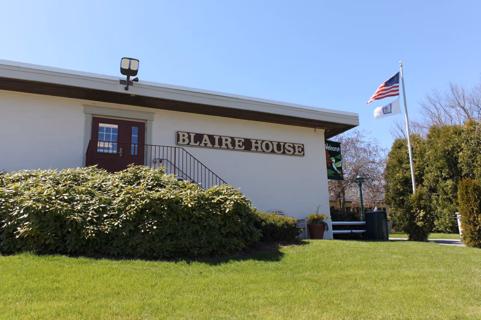 Exterior view of Blaire House building with a white facade, a sign reading 'BLAIRE HOUSE', a set of stairs leading to a door, green bushes in front, and two flags on a flagpole against a clear blue sky.
