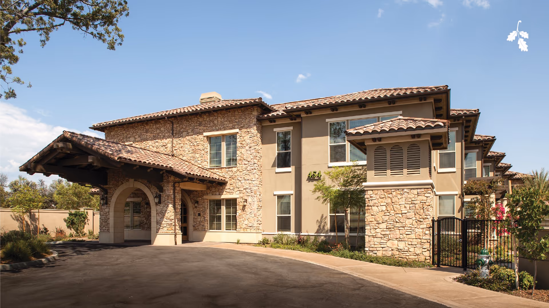 Exterior view of a two-story building with stone and stucco facade, tiled roof, and an arched entrance canopy. The building is surrounded by landscaping and a paved driveway under a clear blue sky.